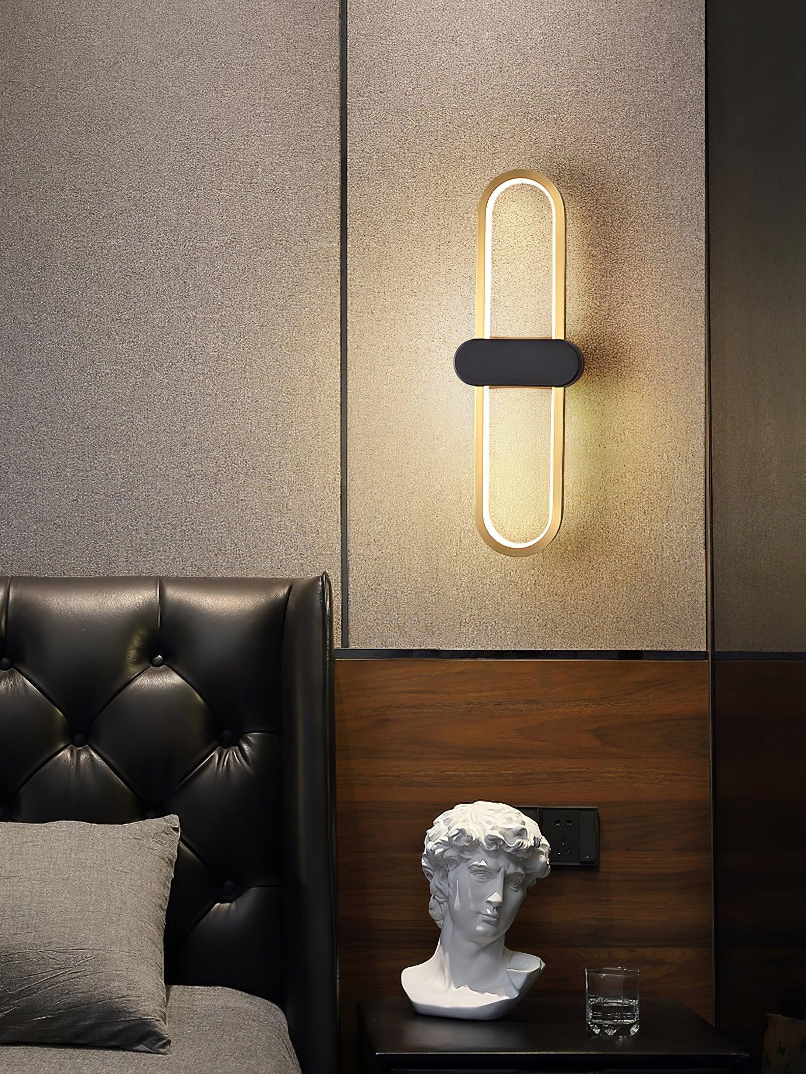 Gold-loop and black-centre wall lamp beside a black tufted headboard above a white head ornament