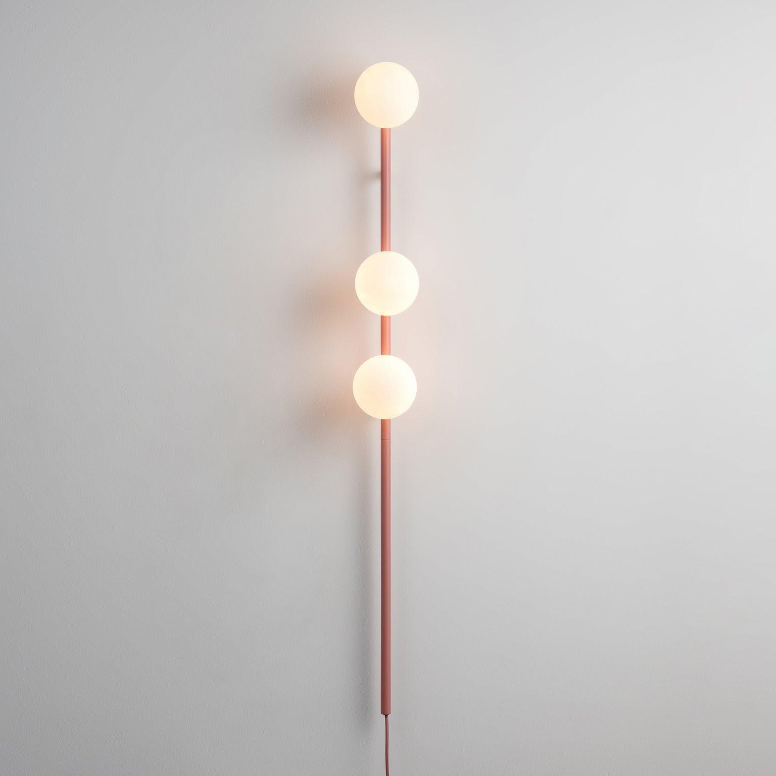 Pink-stem wall light illuminated on pale wall, three white globes, long lower rod and matching pink cord