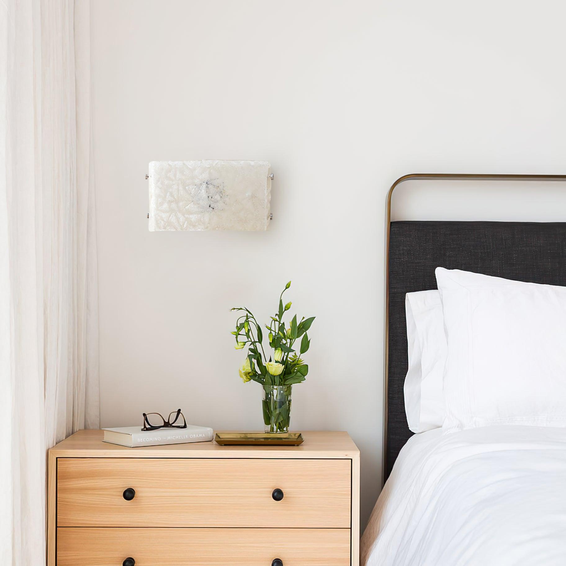 Unlit white glass wall light beside a black brass-edged headboard, above a wood cabinet with flowers and books
