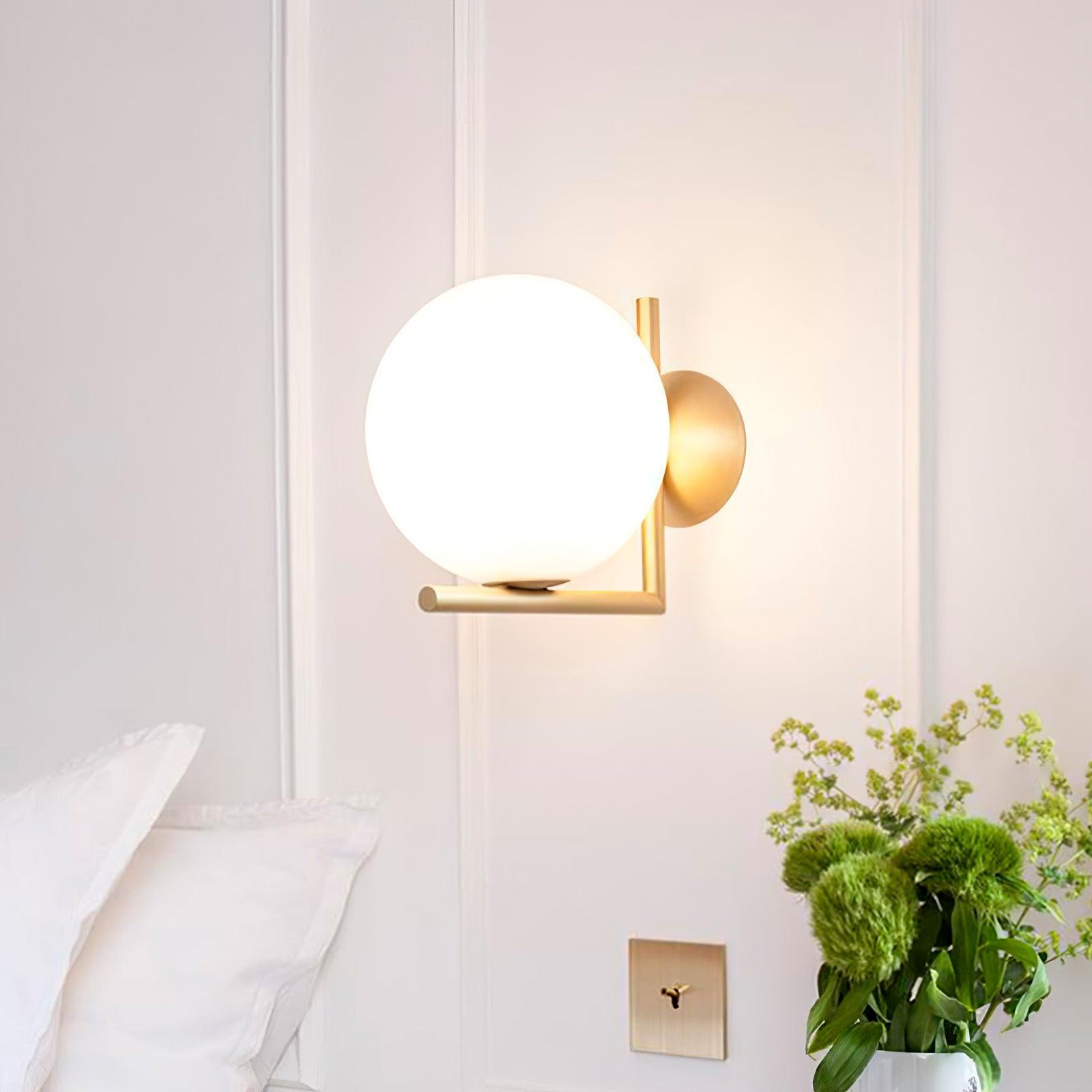 Illuminated white globe wall light with a gold-tone frame beside bedding and flowers