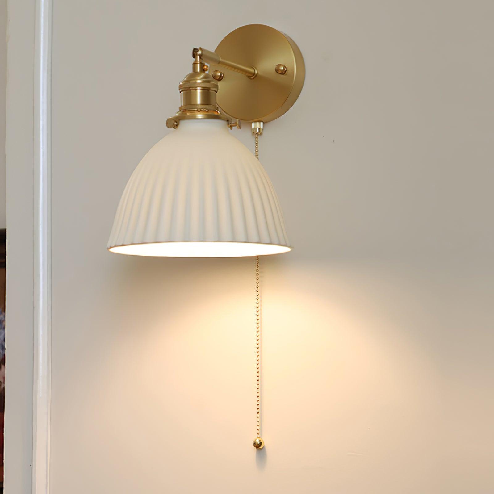 Close front-oblique view of a lit pleated ceramic shade, brass holder, round plate and round-ended chain