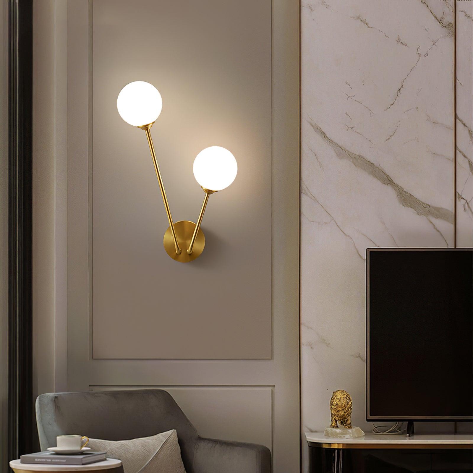 White two-globe wall light above a grey chair beside a television and marble-pattern wall panel