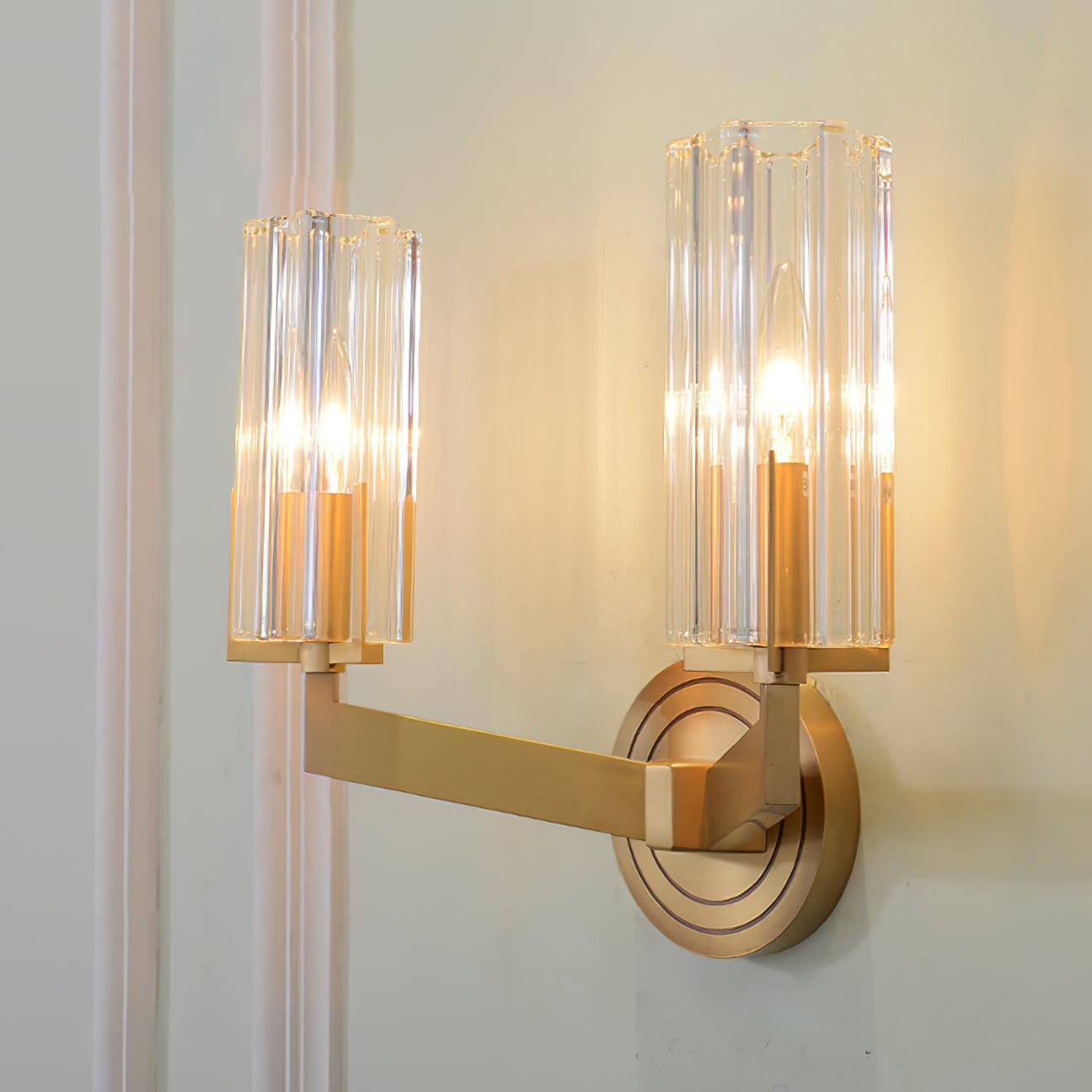 Oblique lit twin-shade Kayla on pale wall beside trim, showing fluted glass cylinders, brass angular arms and ringed plate
