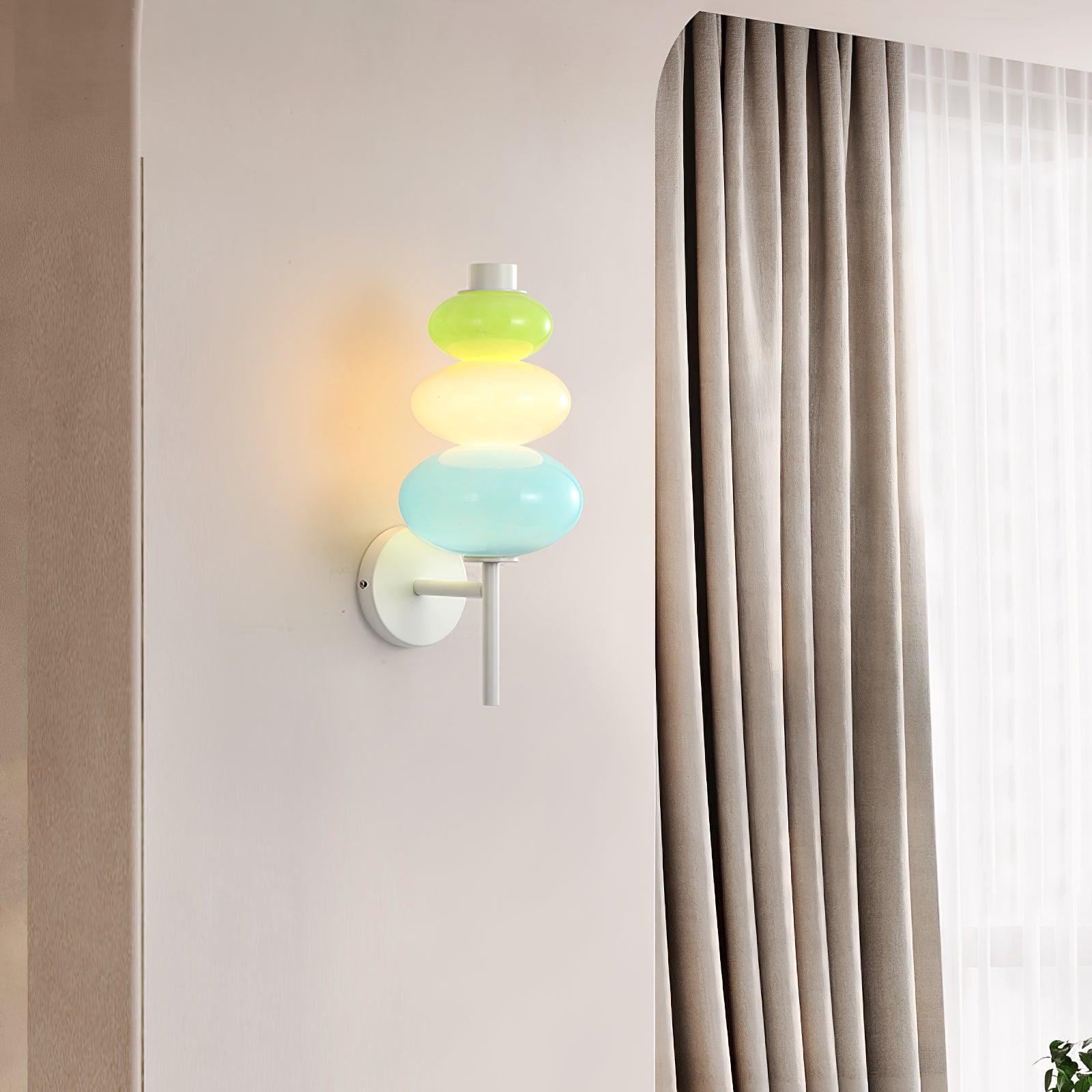 Lit multicolored glass wall light beside white curtains, seen with its wall plate on the left