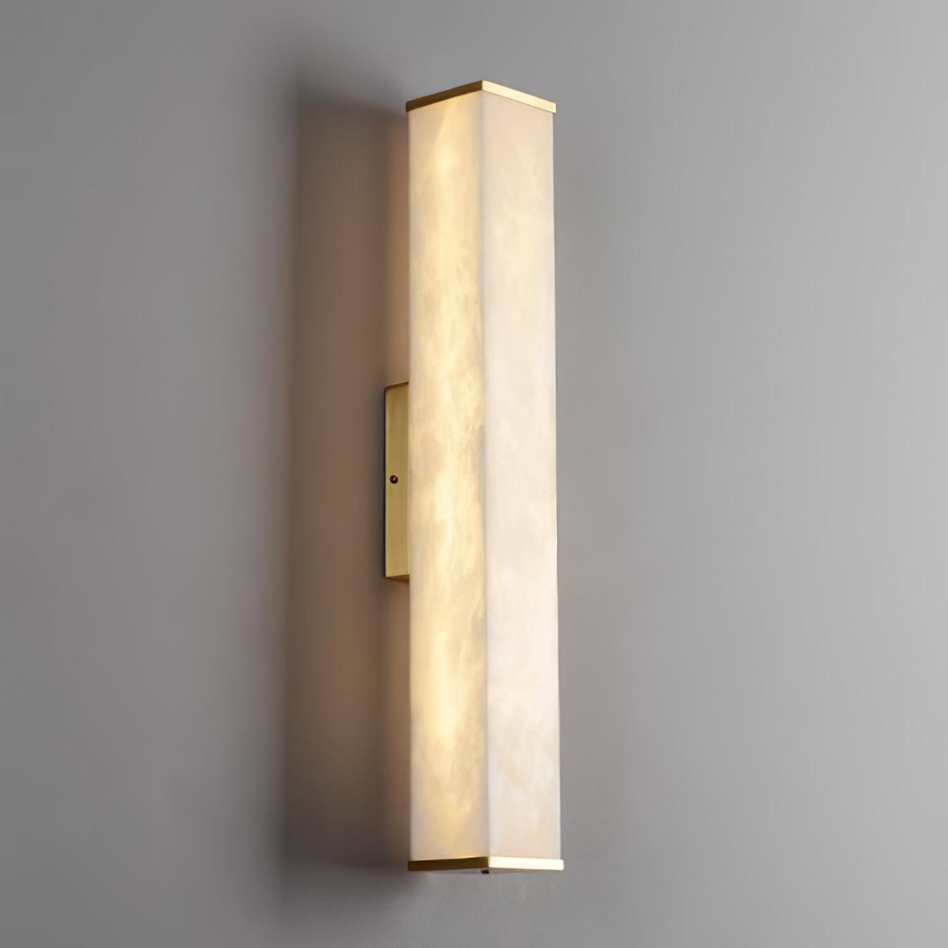 Illuminated rectangular alabaster wall light with thin brass caps, a left-side mounting plate and a soft shadow on a gray wall