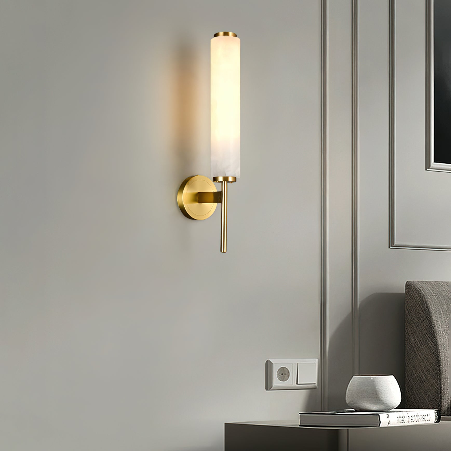 Illuminated white cylinder wall sconce with gold-coloured fittings on a grey wall beside upholstered furniture