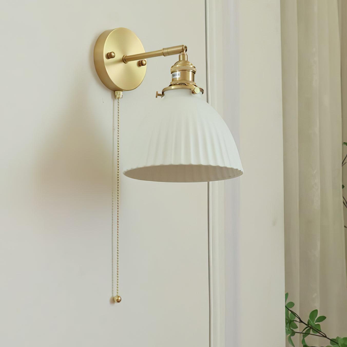 Unlit white pleated shade showing its lower edge and interior, with brass fittings and a hanging chain beside a curtain