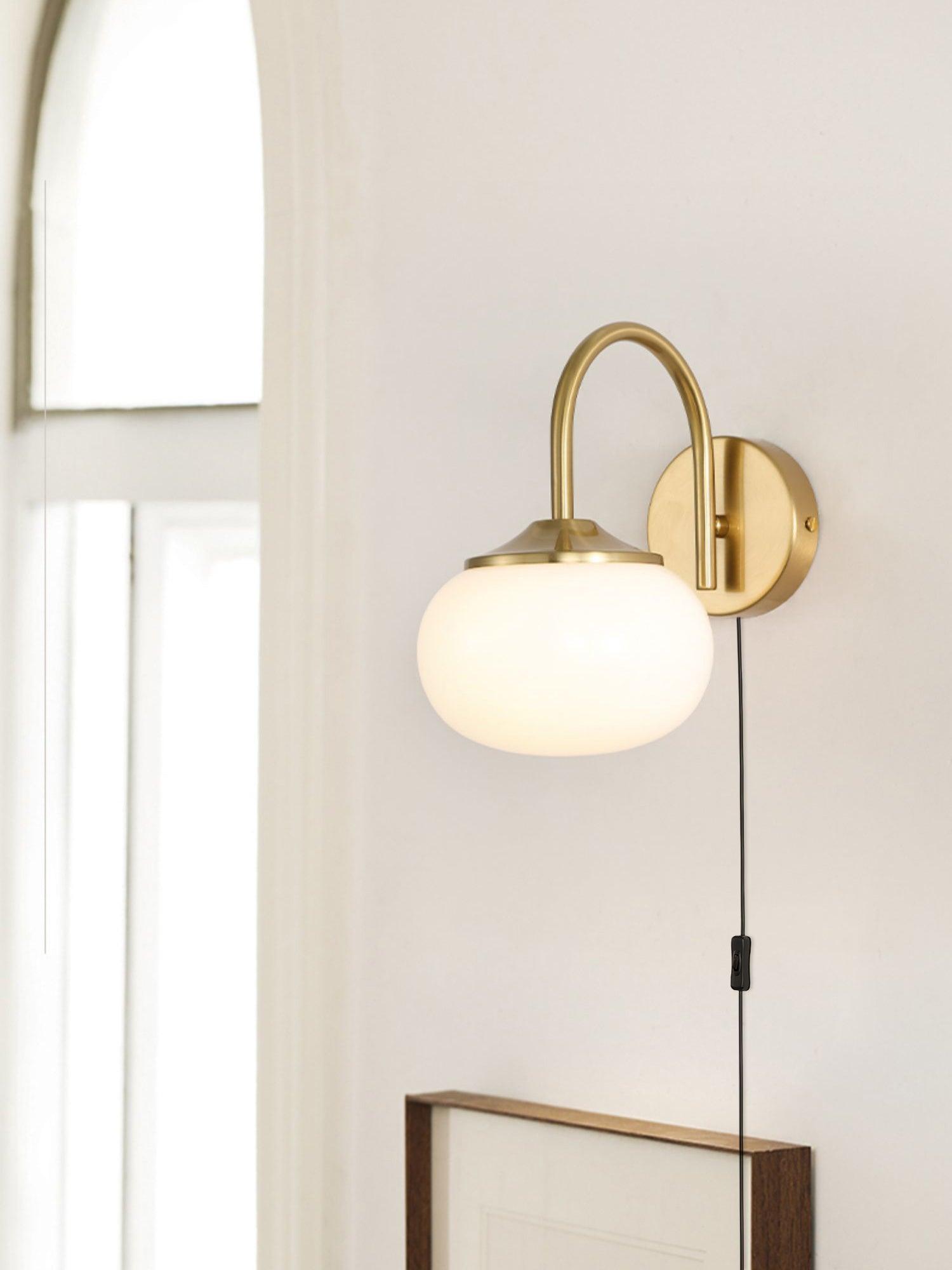 Pale glass wall sconce beside a bright window above a framed picture, with gold-tone fittings and hanging black cord switch