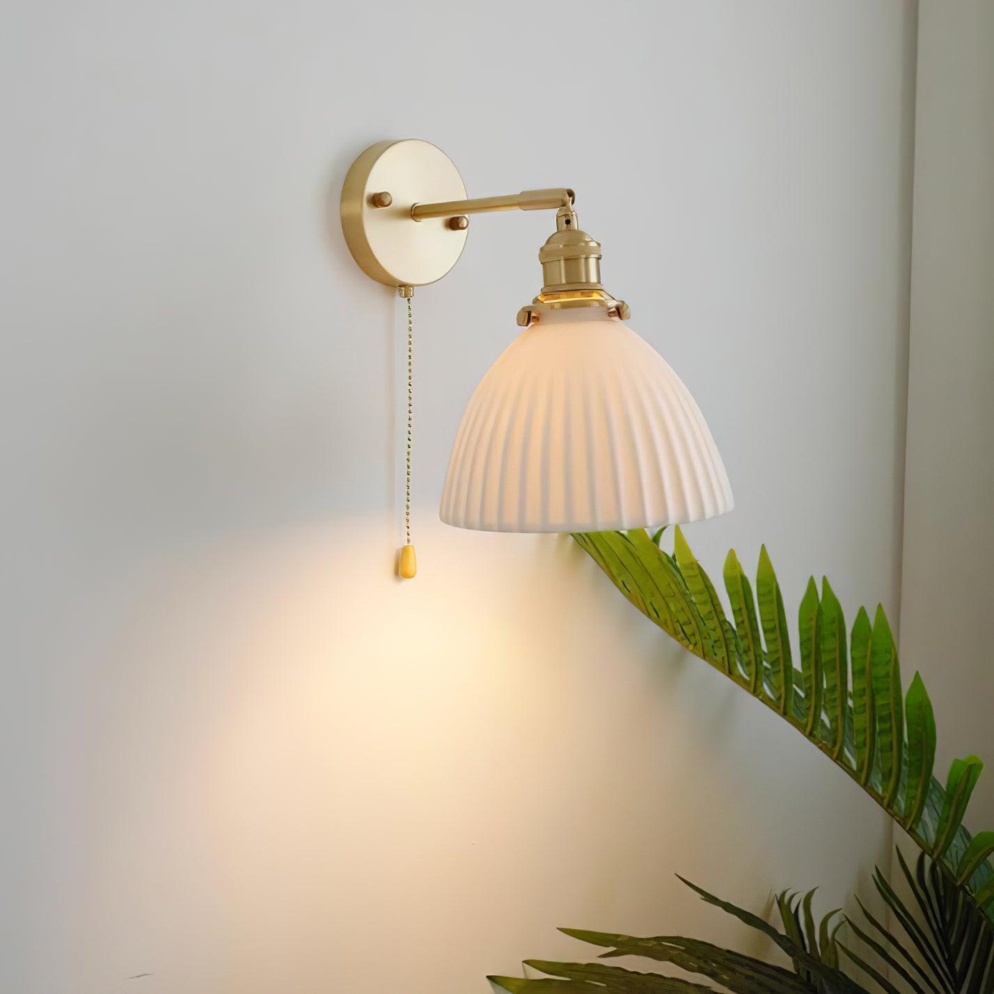 Glowing white pleated shade above green leaves, with straight brass arm and a teardrop-ended chain