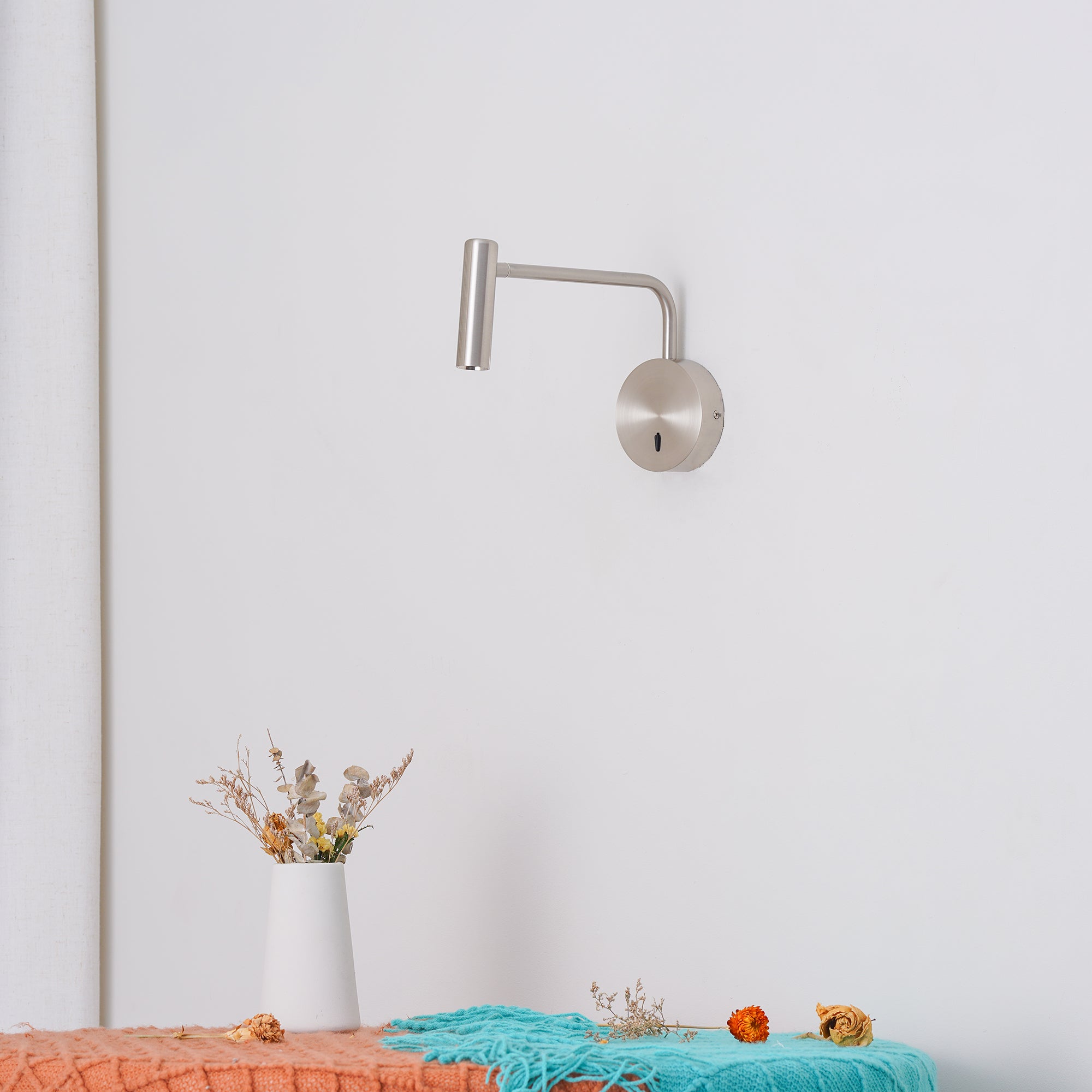 Brushed metal wall light above a flower vase and orange and turquoise draped textiles