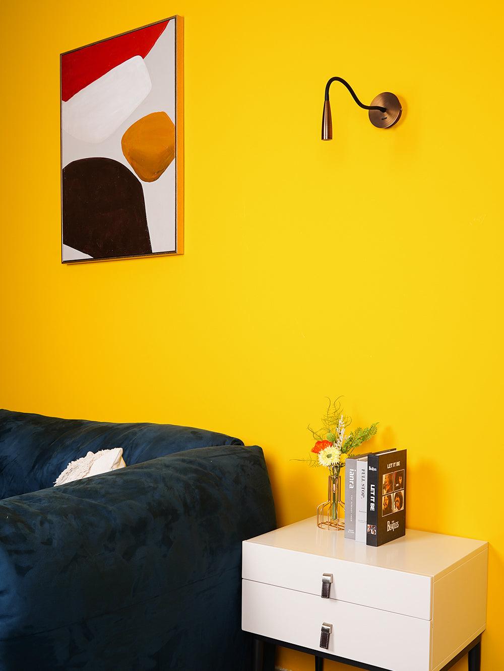 Bronze flexible-arm wall light above white drawer cabinet beside blue sofa and abstract print on yellow wall