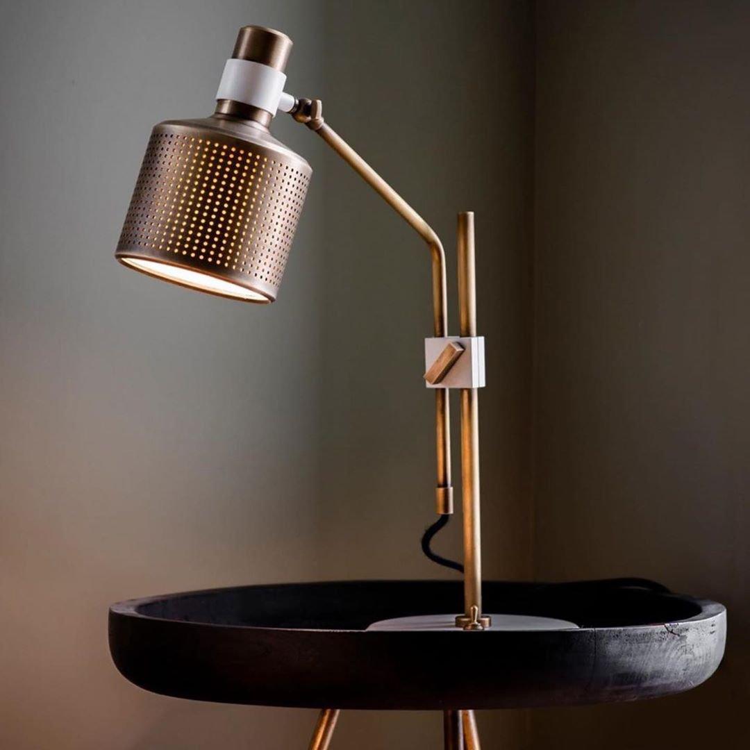 Lit gold-and-white single-head lamp on a black tray table against a grey wall, with a perforated cylindrical shade