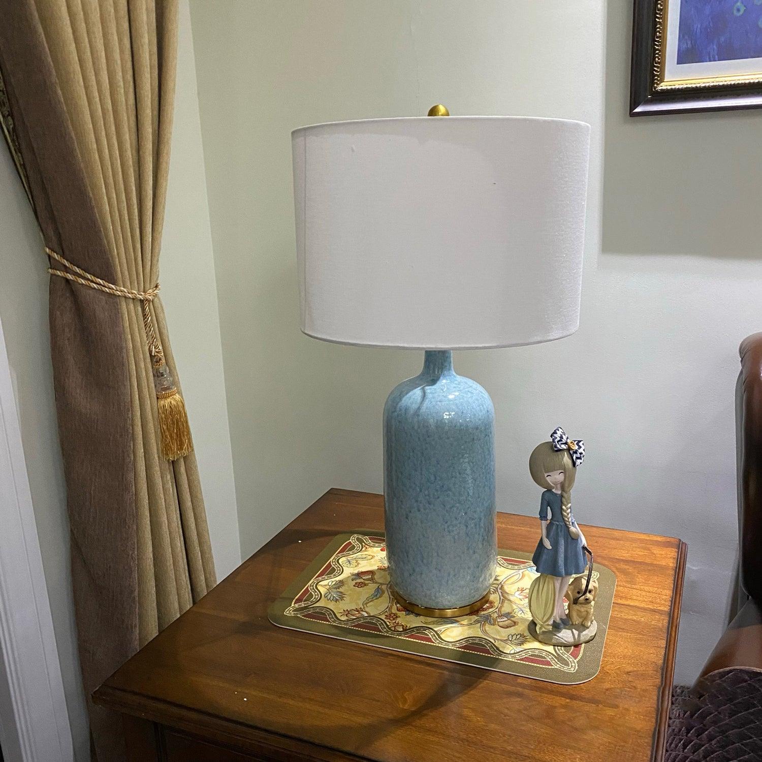 Culloden blue table lamp with a pale drum shade on a wooden table beside a curtain
