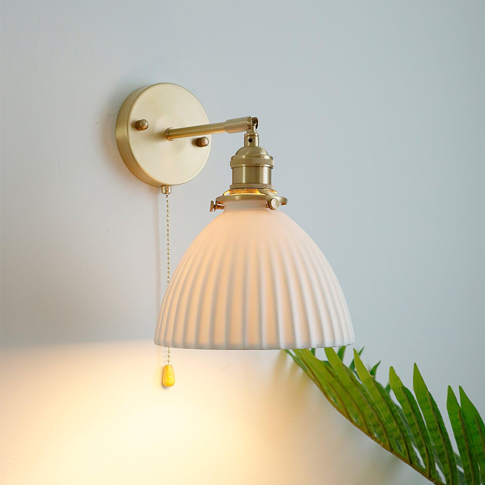Close lit white pleated shade, knurled brass holder and round wall plate beside green fronds