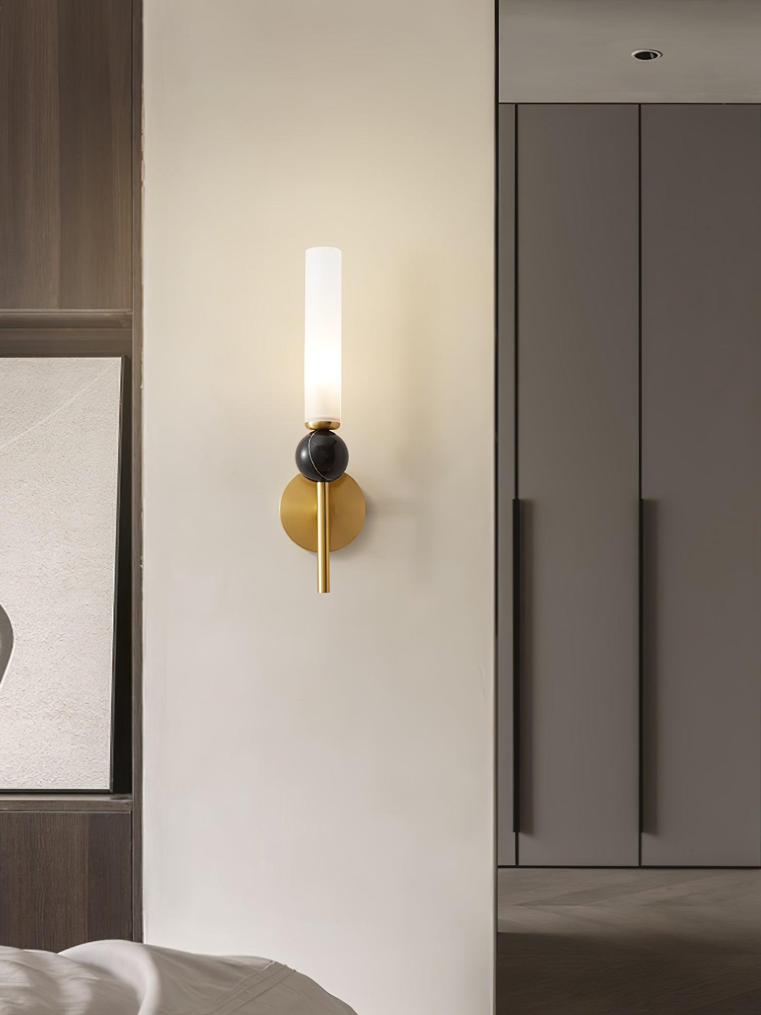 Straight brass wall light with dark veined sphere and lit white cylinder on bedroom partition, beside bed and fitted cabinets