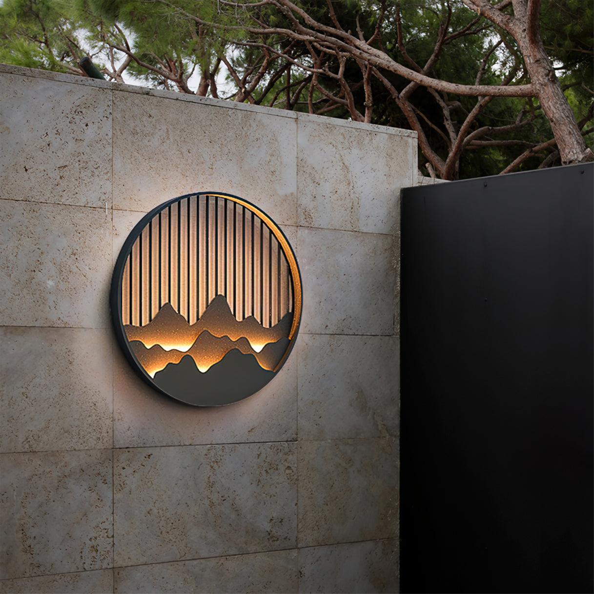 Lit circular Mountain wall light on pale tiled garden wall beside dark gate, with tree branches overhead