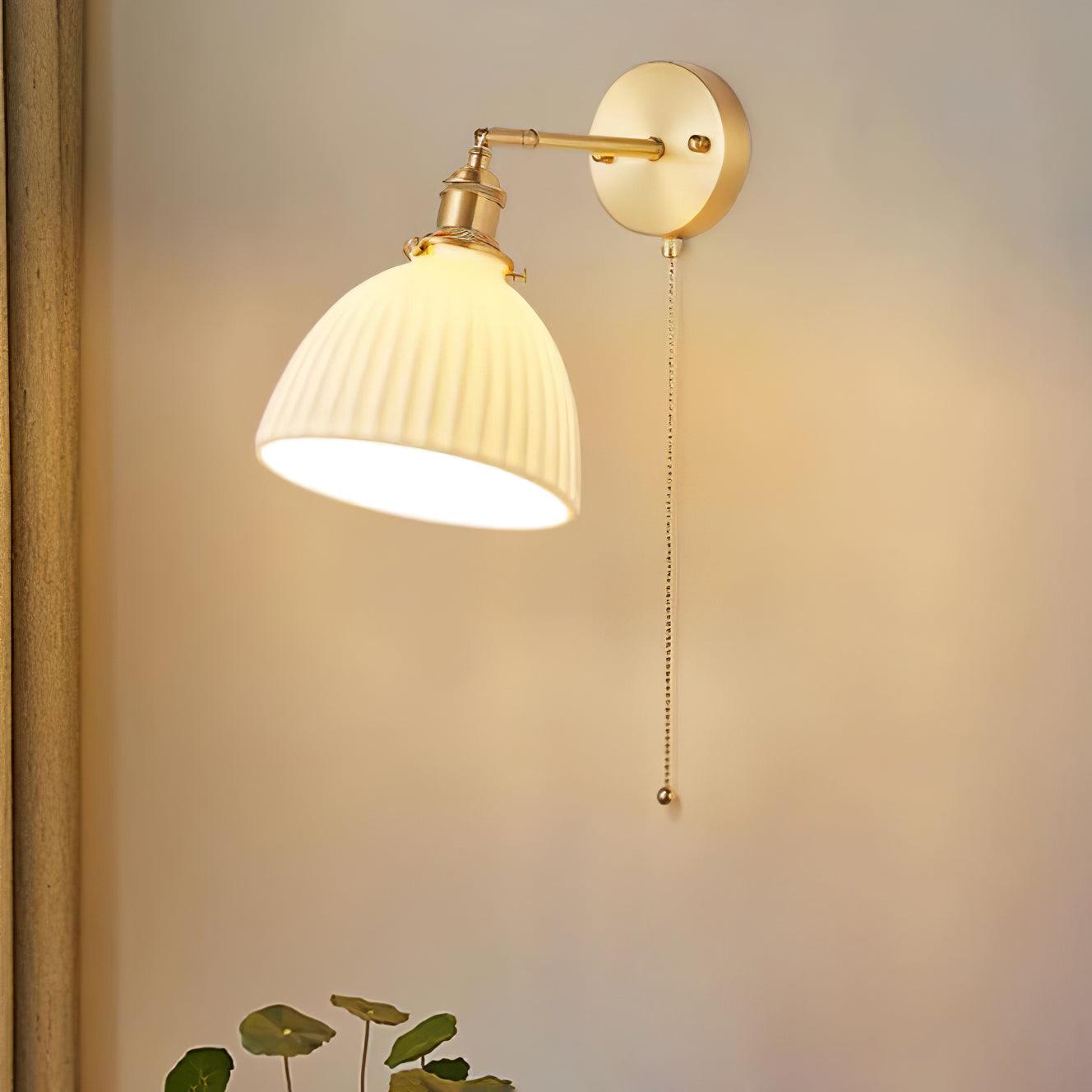 Lit ceramic shade tilted down-left beside a curtain and plant, with brass wall plate and a long hanging chain