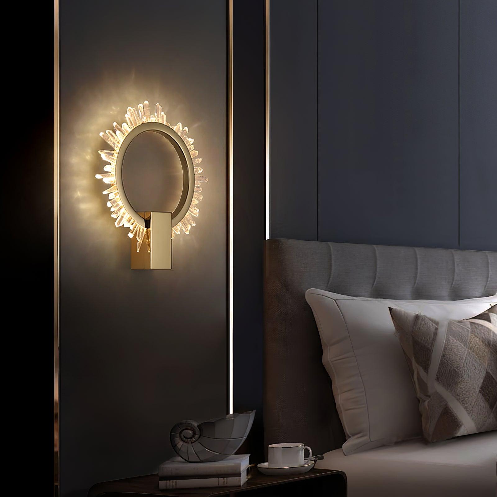 Lit crystal-ring wall light beside a grey headboard above books, a shell ornament and a cup