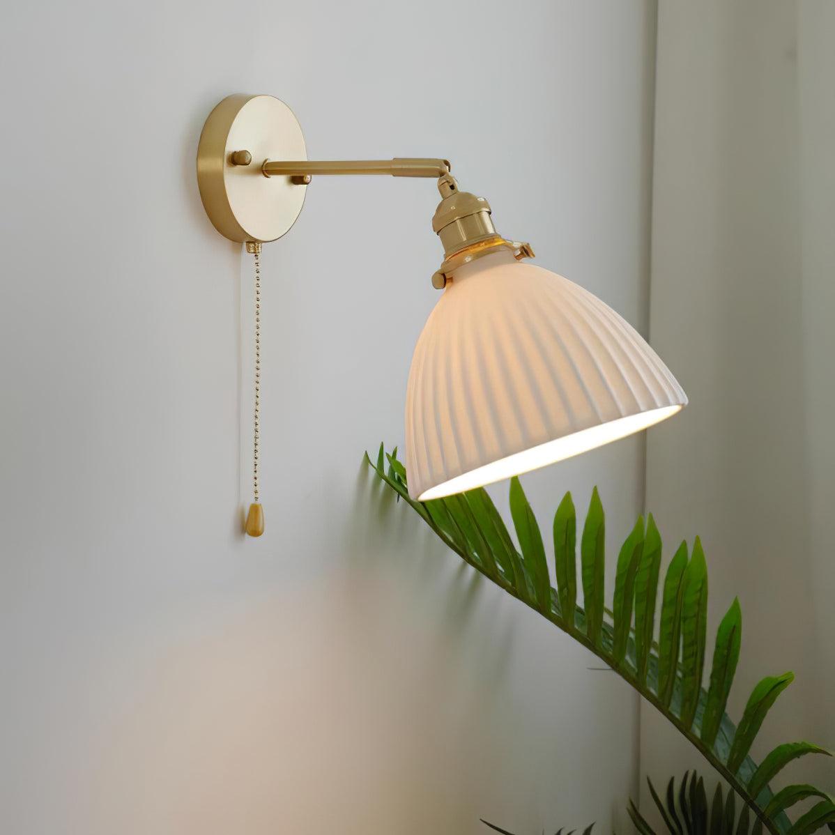 Lit white pleated shade tilted down-right above green fronds, on a brass arm with a round wall plate