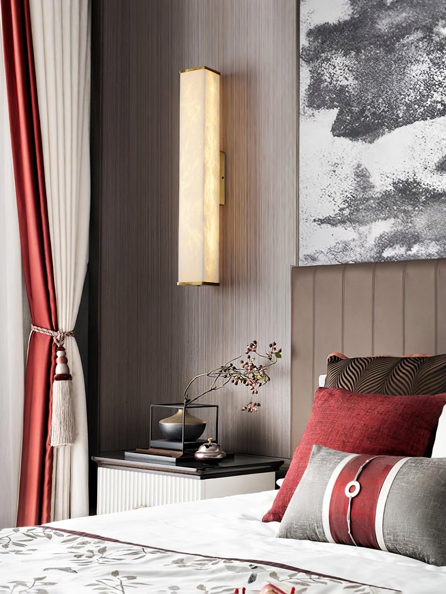 Bedroom view of an alabaster wall light above a bedside table, with red cushions, embroidered bedding and tied curtains