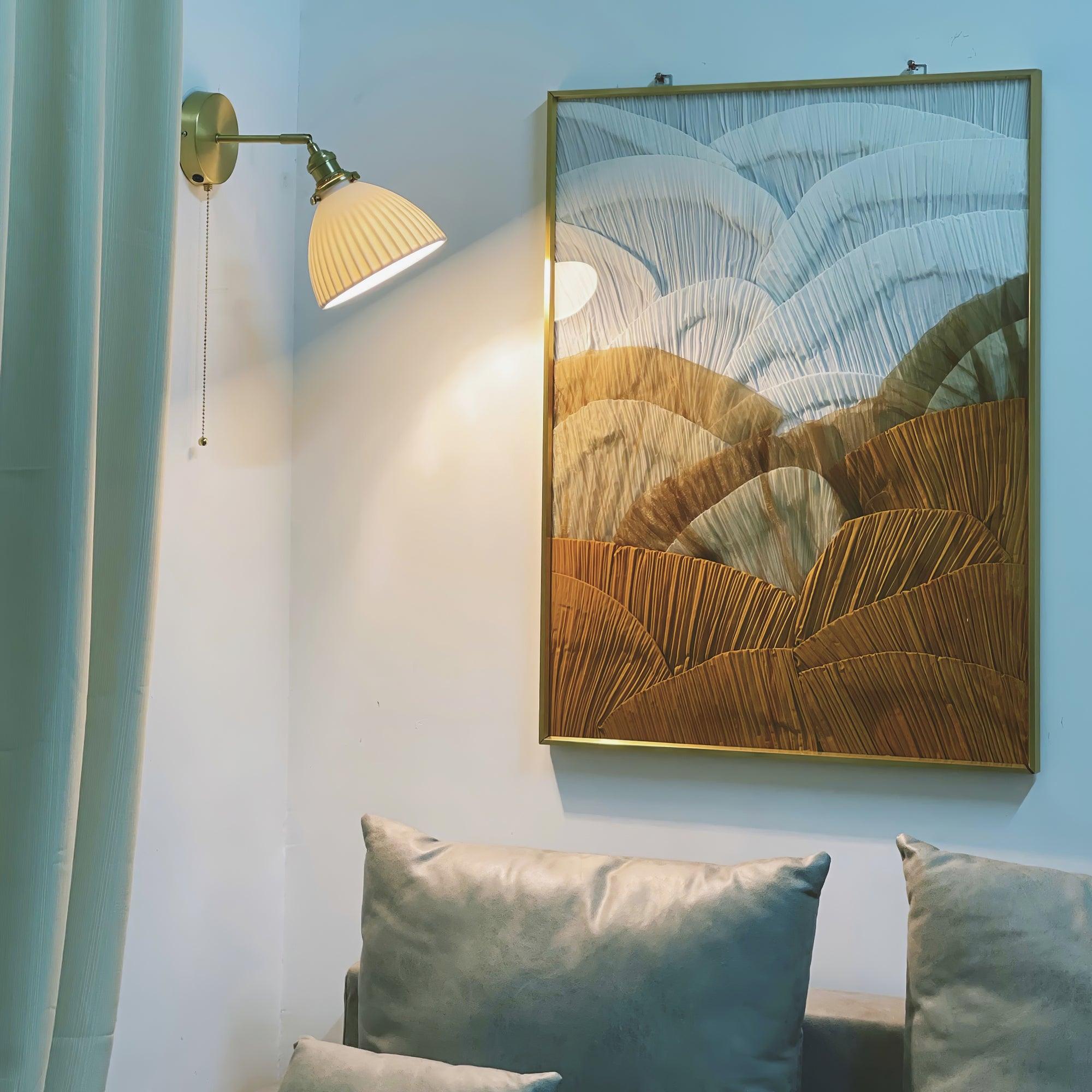 Lit pleated wall lamp beside a large gold-framed abstract picture above sofa cushions