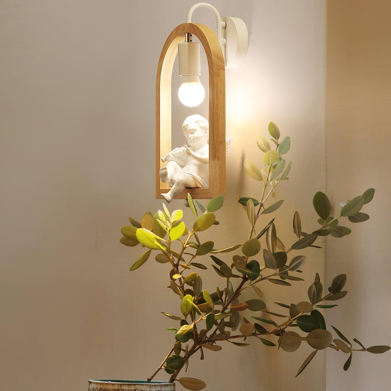 Oblique view of the cherub wall light beside a leafy plant, showing the violin ornament, wooden arch and white wallplate
