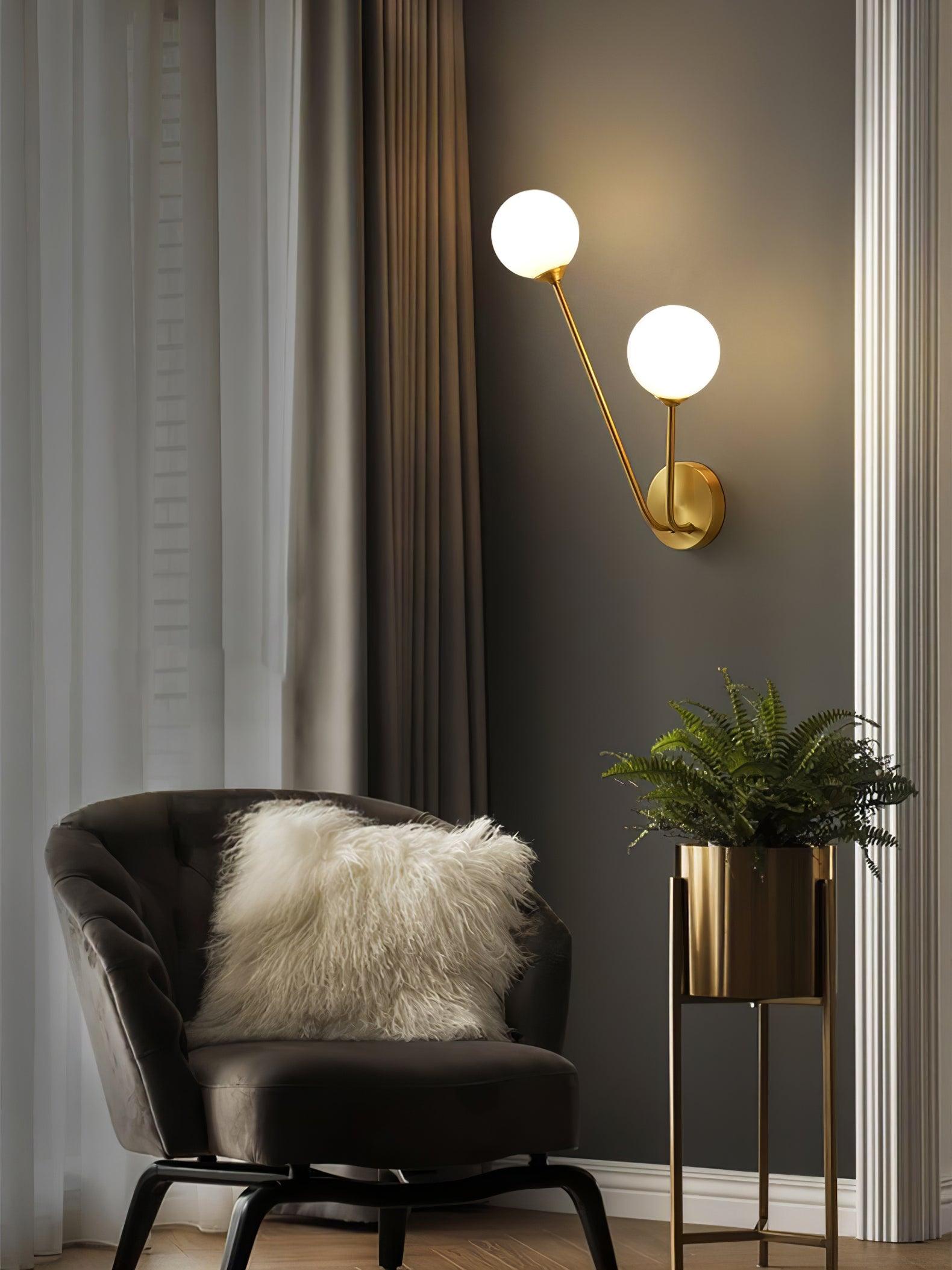White globe wall light above a dark chair and white cushion, beside curtains and a fern in a gold-tone planter