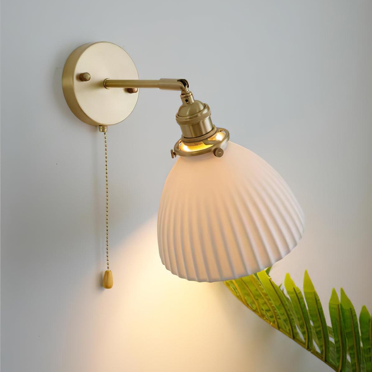 Close lit ceramic shade tilted diagonally beside green leaves, with a brass arm and teardrop-ended chain