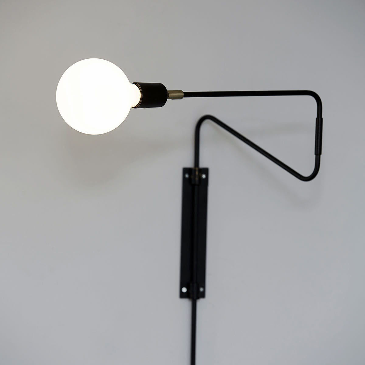 Black bent-arm wall lamp with a glowing white globe bulb and a narrow wallplate