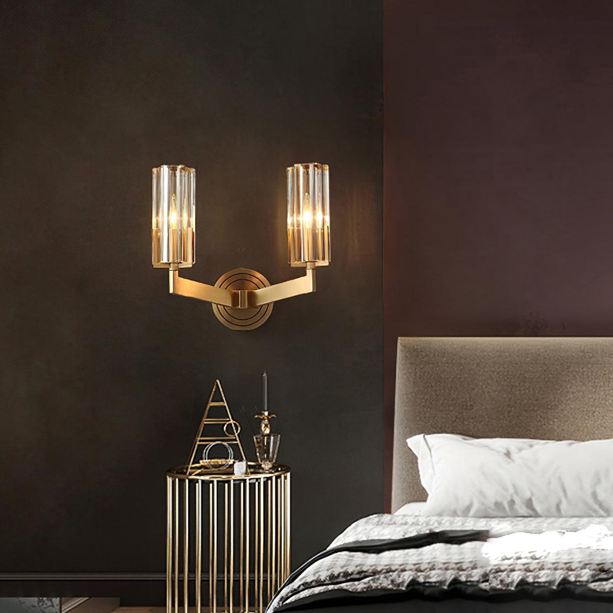 Lit twin-shade Kayla beside upholstered bed and slim gold-tone table holding triangular ornament and candle