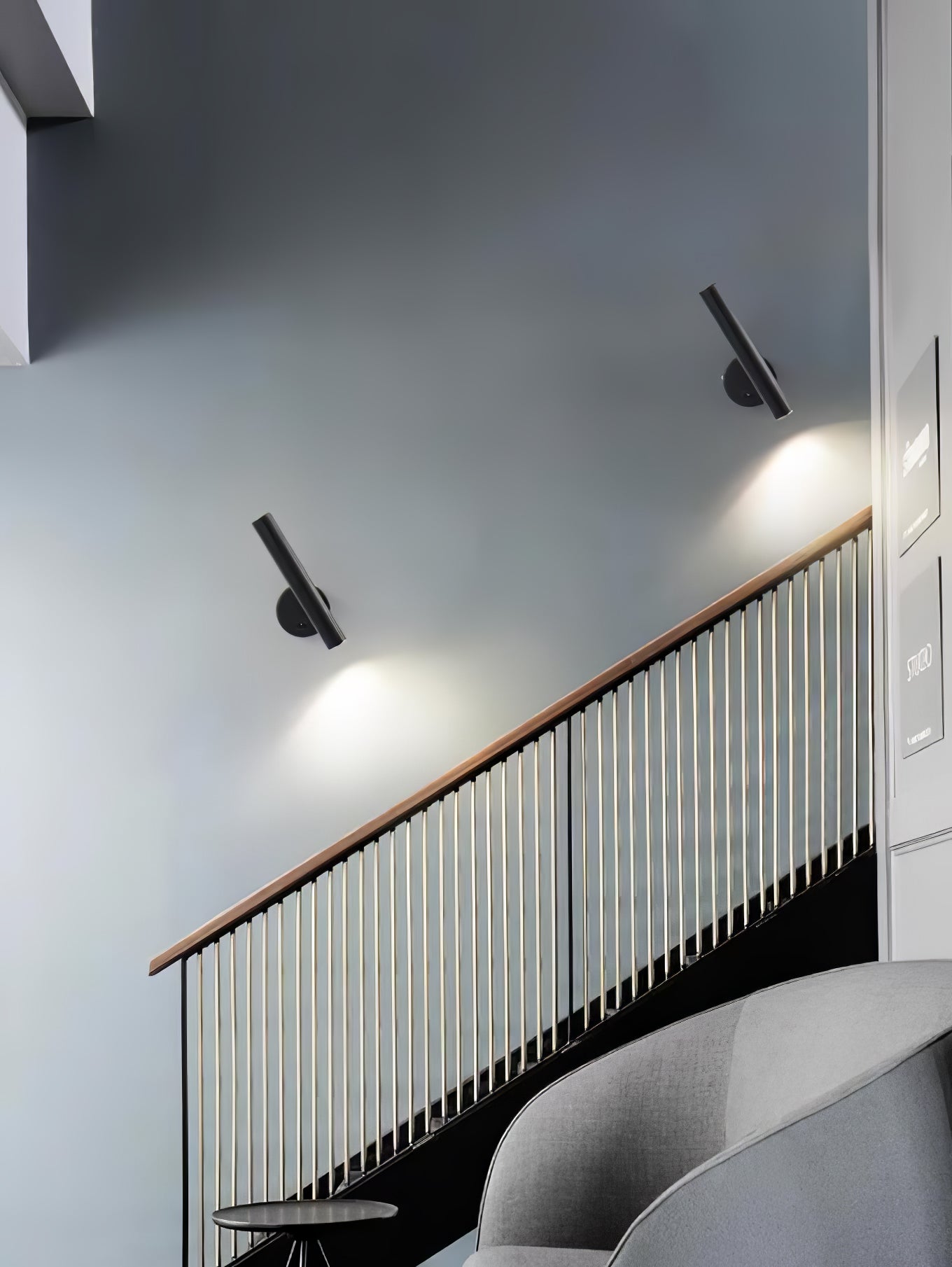 Wider view of two black tubular wall lights on a stair wall above a wood-topped metal railing