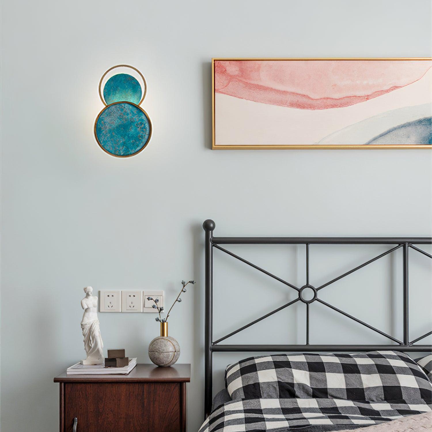 Turquoise Satellite light above a wooden bedside cabinet beside a black metal bed frame and pink-blue framed artwork.
