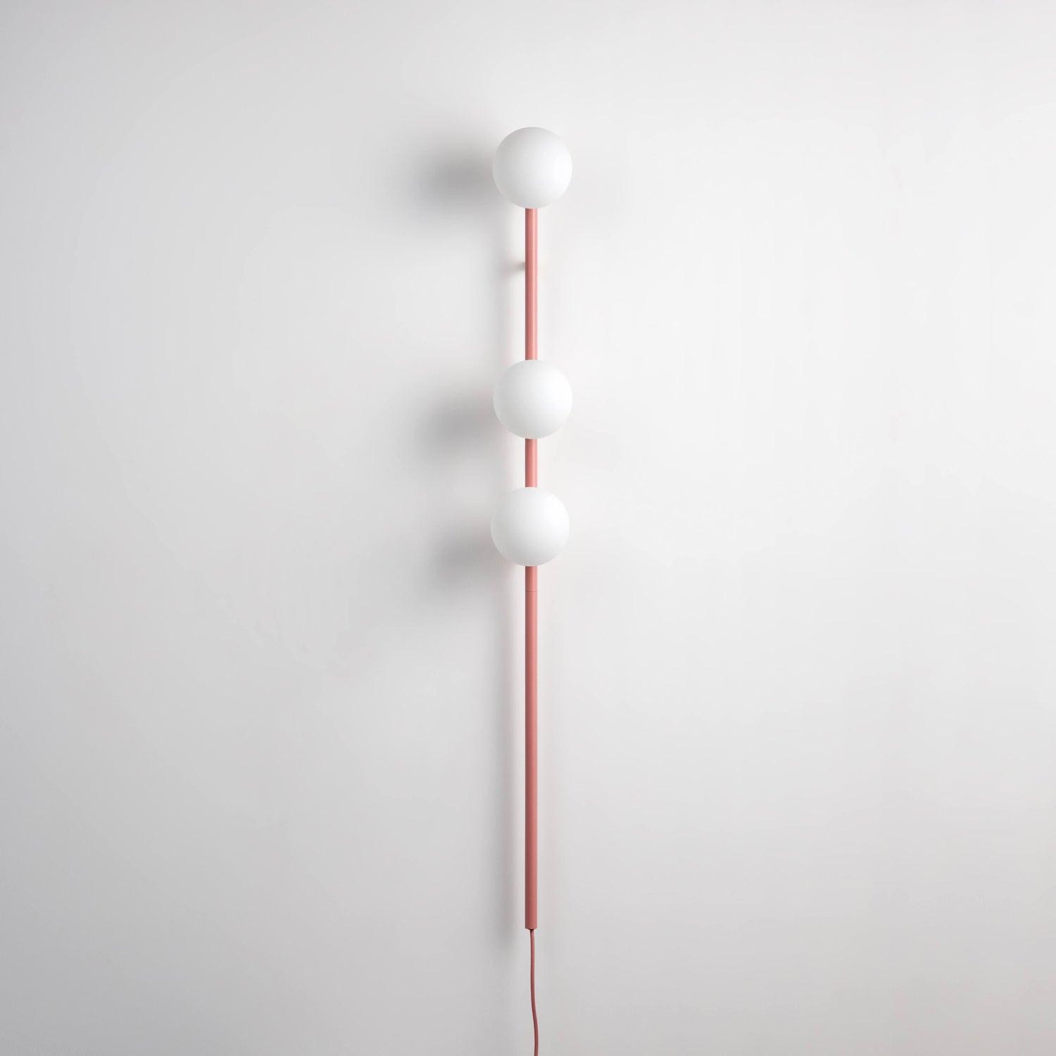 Unlit pink wall light against pale wall, three white glass globes, slender stem and pink lower cable