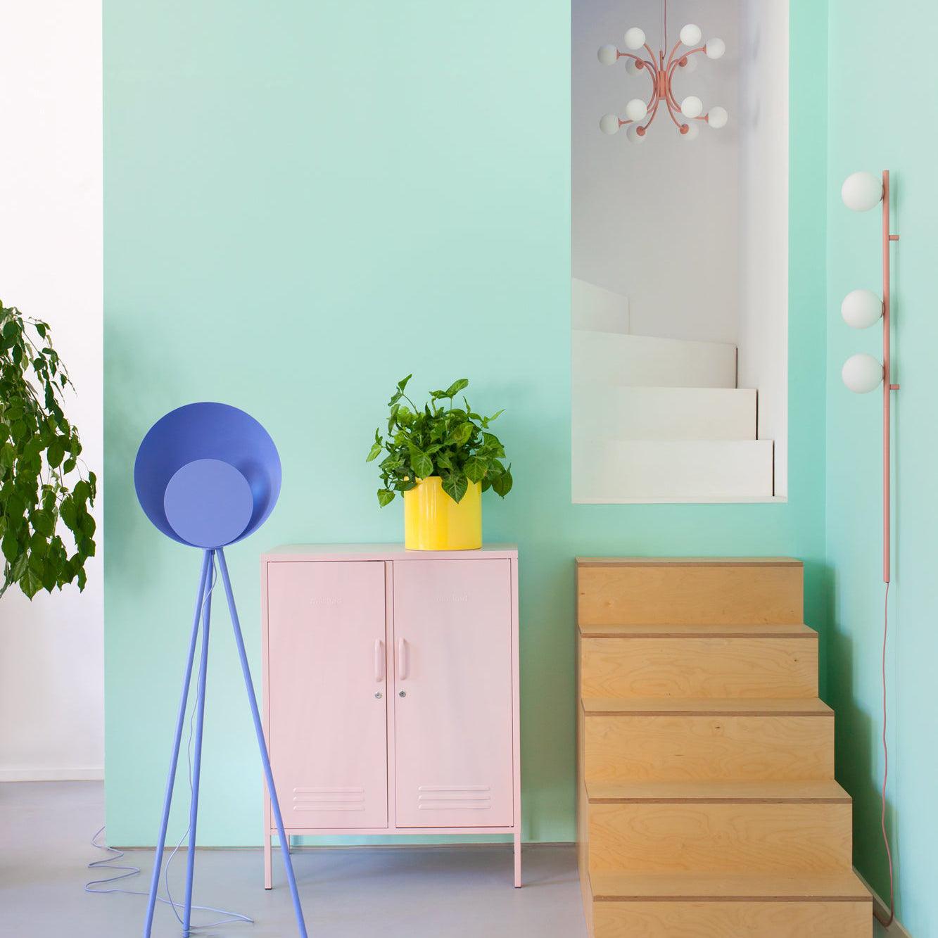 Pink three-globe wall light in mint room by wooden steps, pink cabinet and mirror, with blue tripod lamp nearby