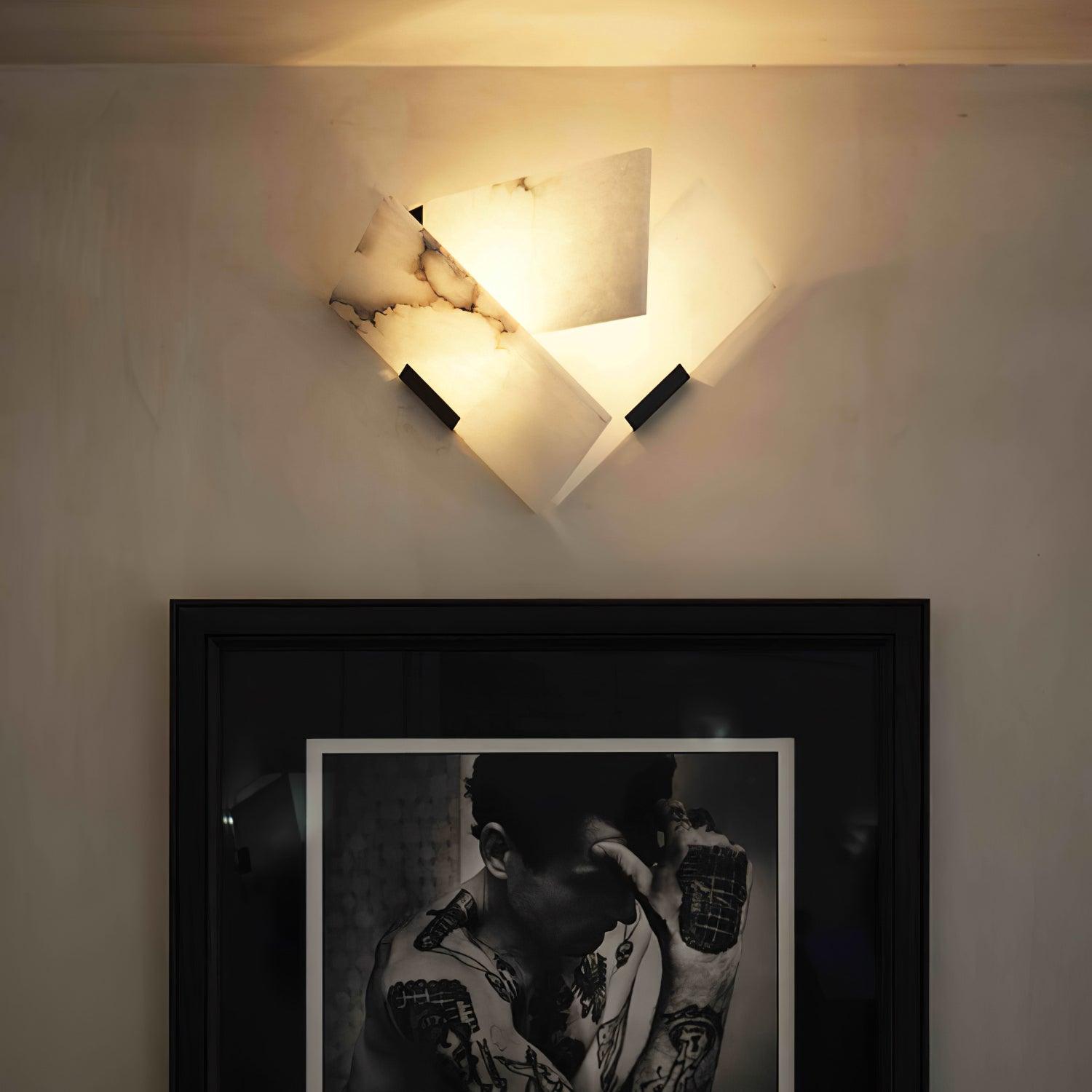 Illuminated angular stone sconce on a pale wall above a black-framed portrait