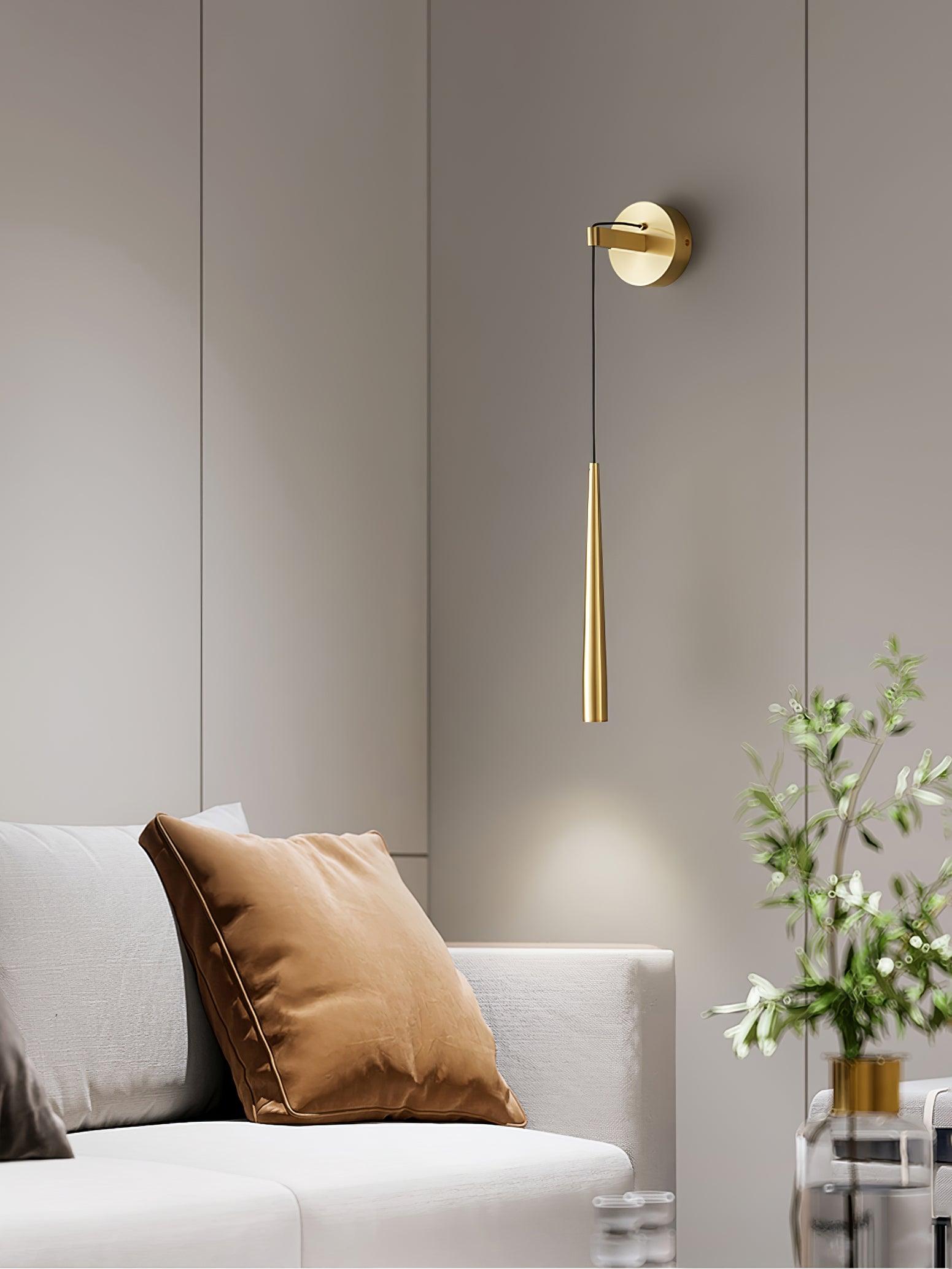 Brass-tone plain-ended wall light above a sofa cushion beside a glass vase of branches