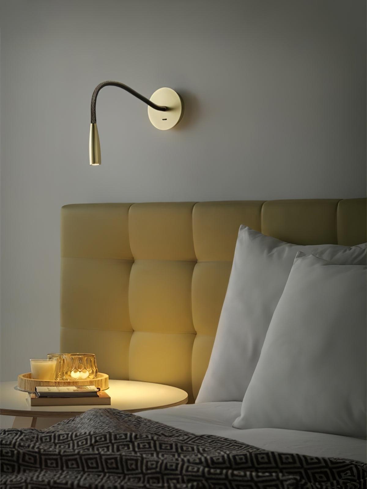 Pale gold-tone wall light with dark textured arm above olive headboard and round bedside table with books and drinks