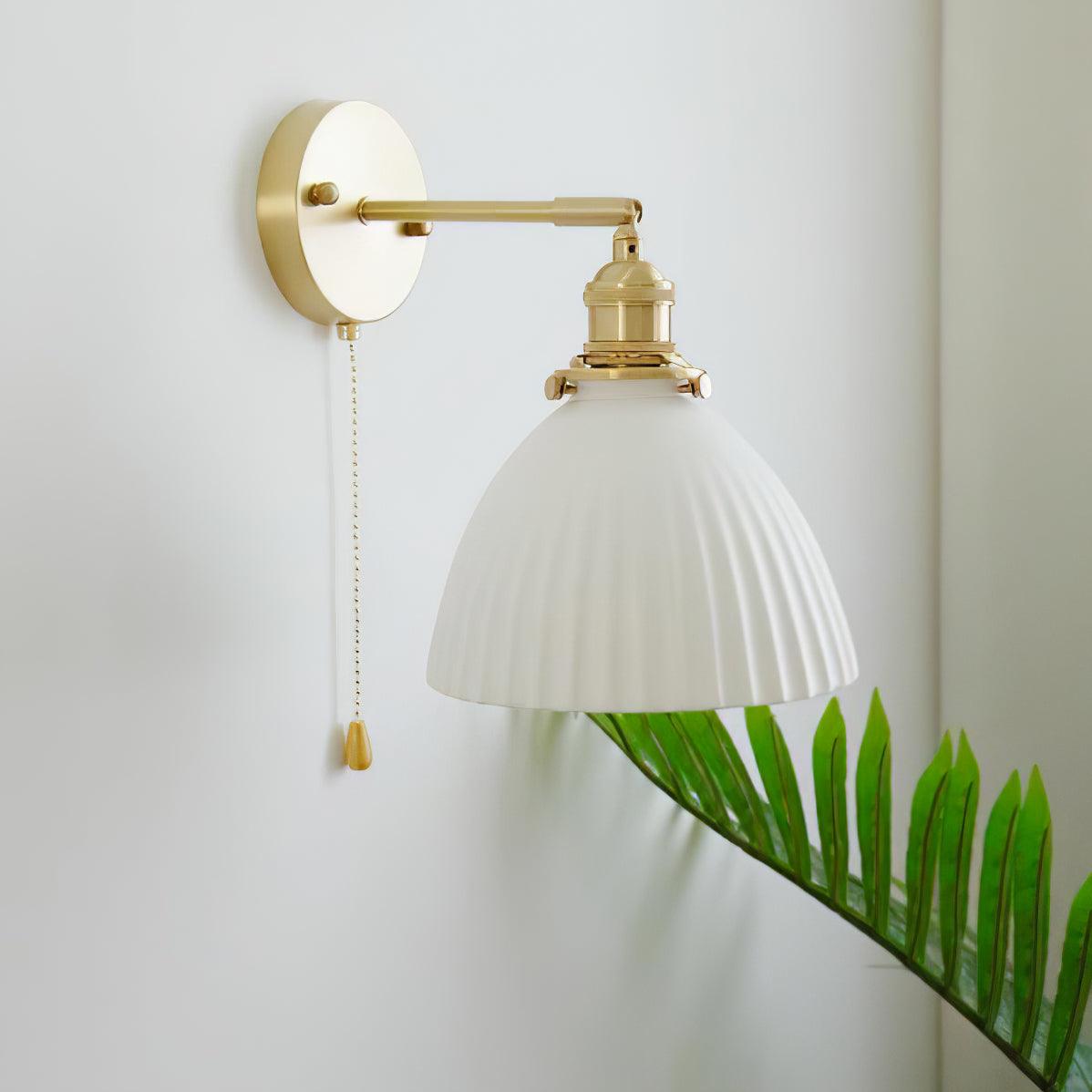 Unlit white pleated ceramic shade below a brass holder, with straight arm, round wall plate and hanging chain