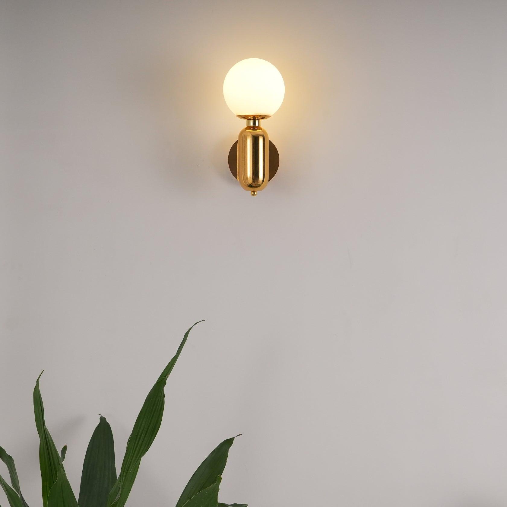 Gold-toned white-globe wall light lit high on pale wall, with large green leaves entering the foreground below