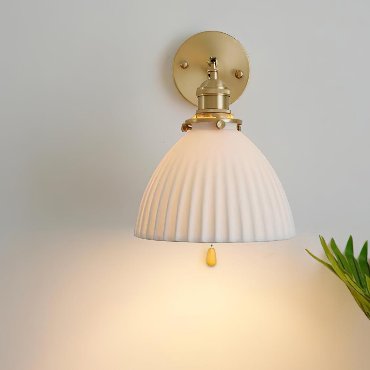 Frontal view of glowing pleated ceramic shade below a brass holder and circular plate, with chain end visible