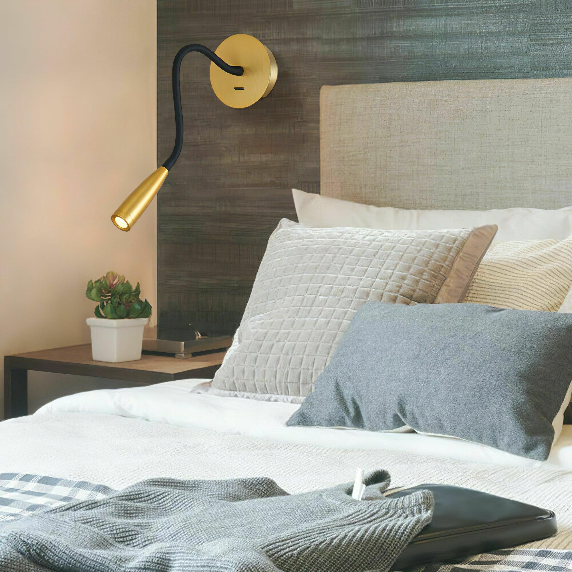 Gold reading light beside grey upholstered headboard, white quilted pillows, blue cushion and bedside plant
