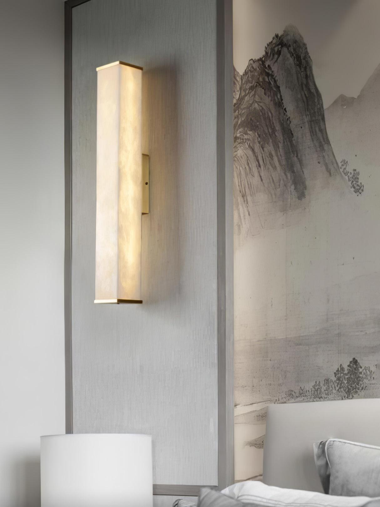 Alabaster wall light with brass ends beside a pale bed and mountain landscape artwork, with a table-lamp shade below