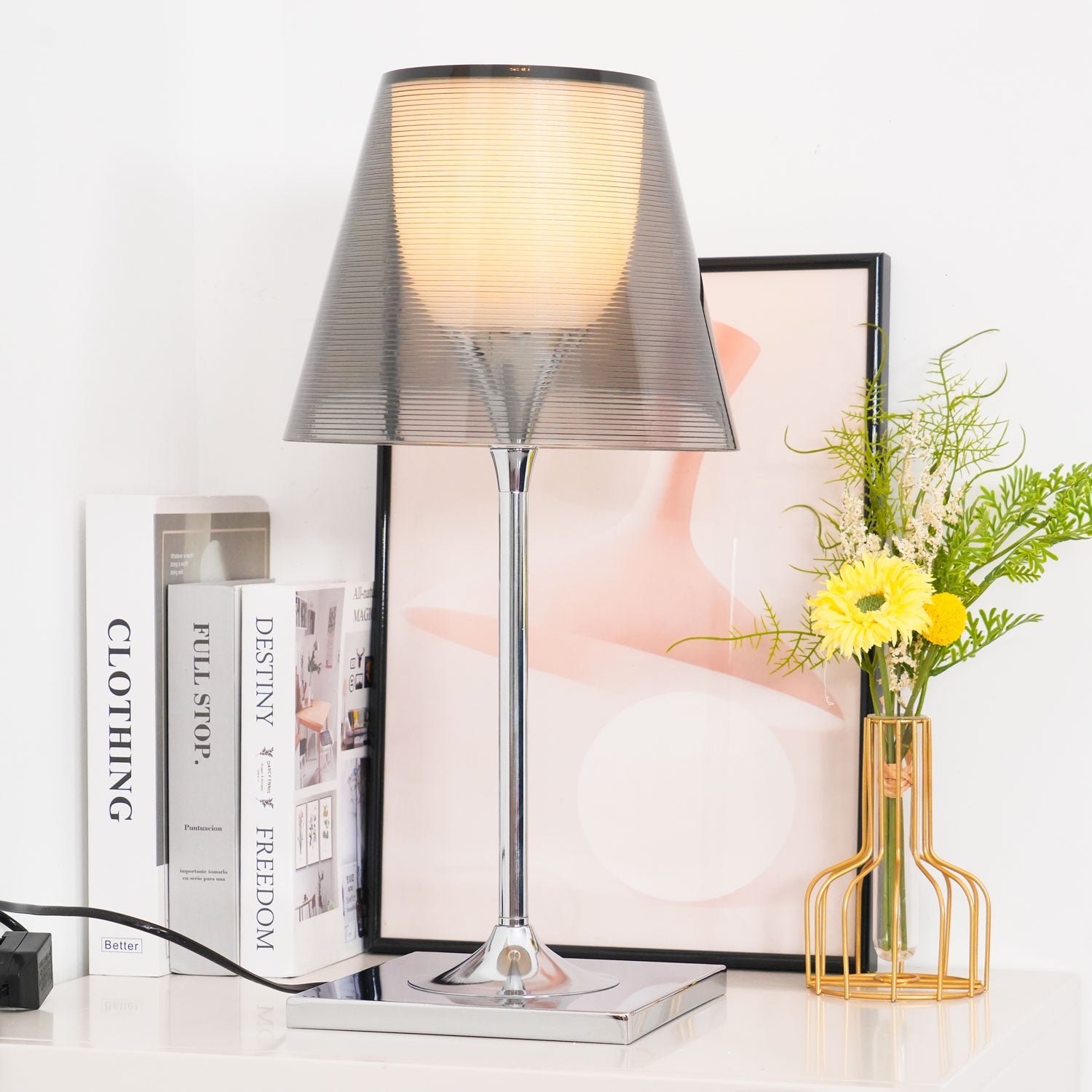 Chrome Prism lamp with a lit smoke-grey shade beside books and framed art