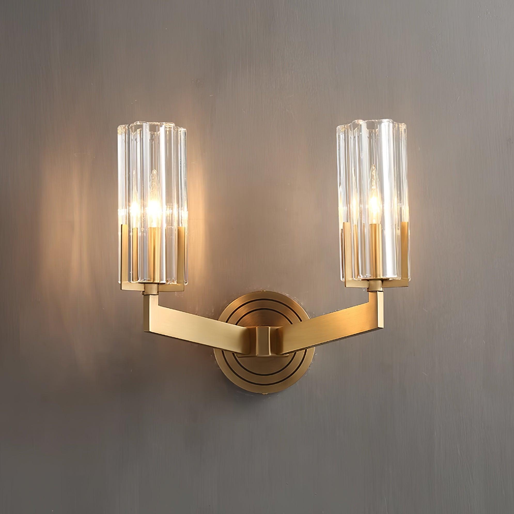 Frontal lit twin clear-glass shades on symmetrical brass arms above concentric-ring wall plate on textured grey wall