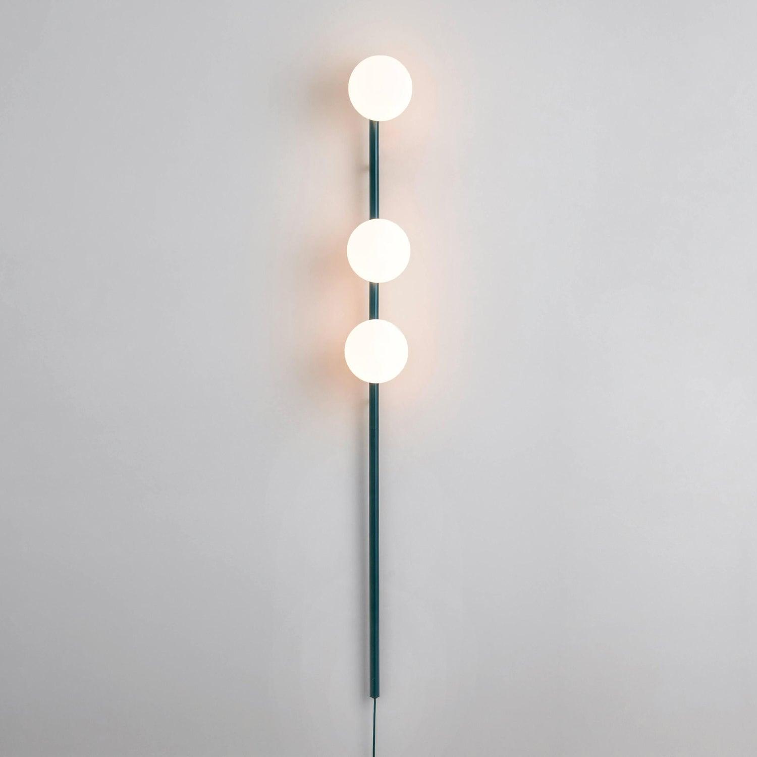 Full lit teal three-globe wall light on pale wall, white glowing spheres and dark lower stem and cable