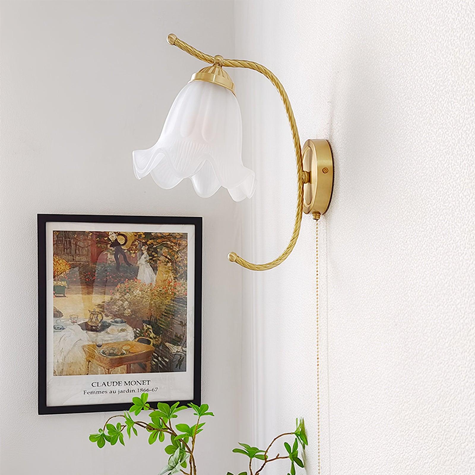 Unlit scalloped glass wall lamp above framed garden artwork and green plant, with twisted brass-finish arm and beaded pull chain
