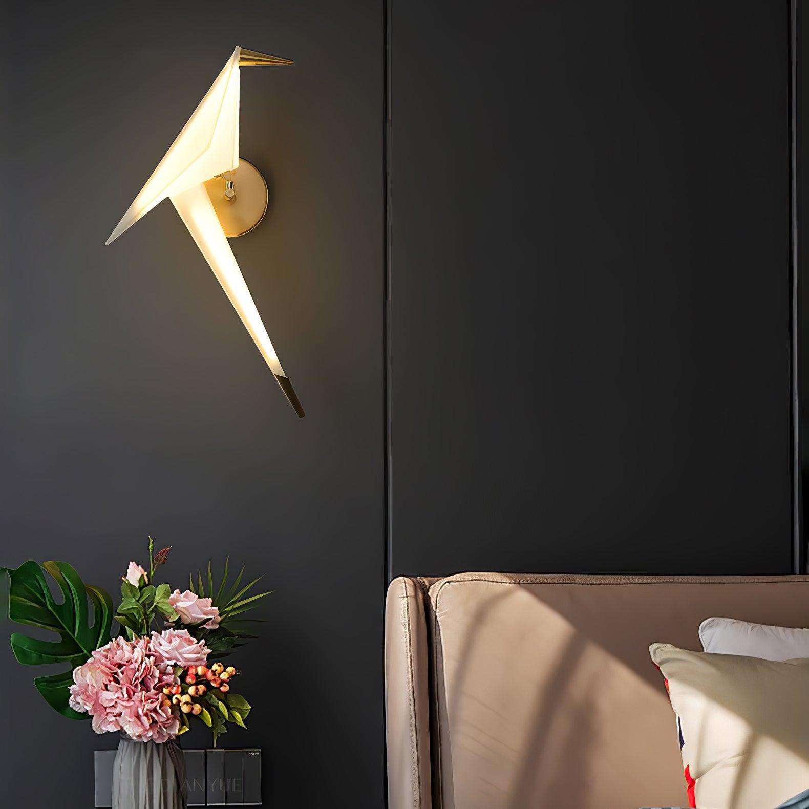 Gold and White bird wall light glowing against a dark wall beside a beige chair and a vase of pink flowers
