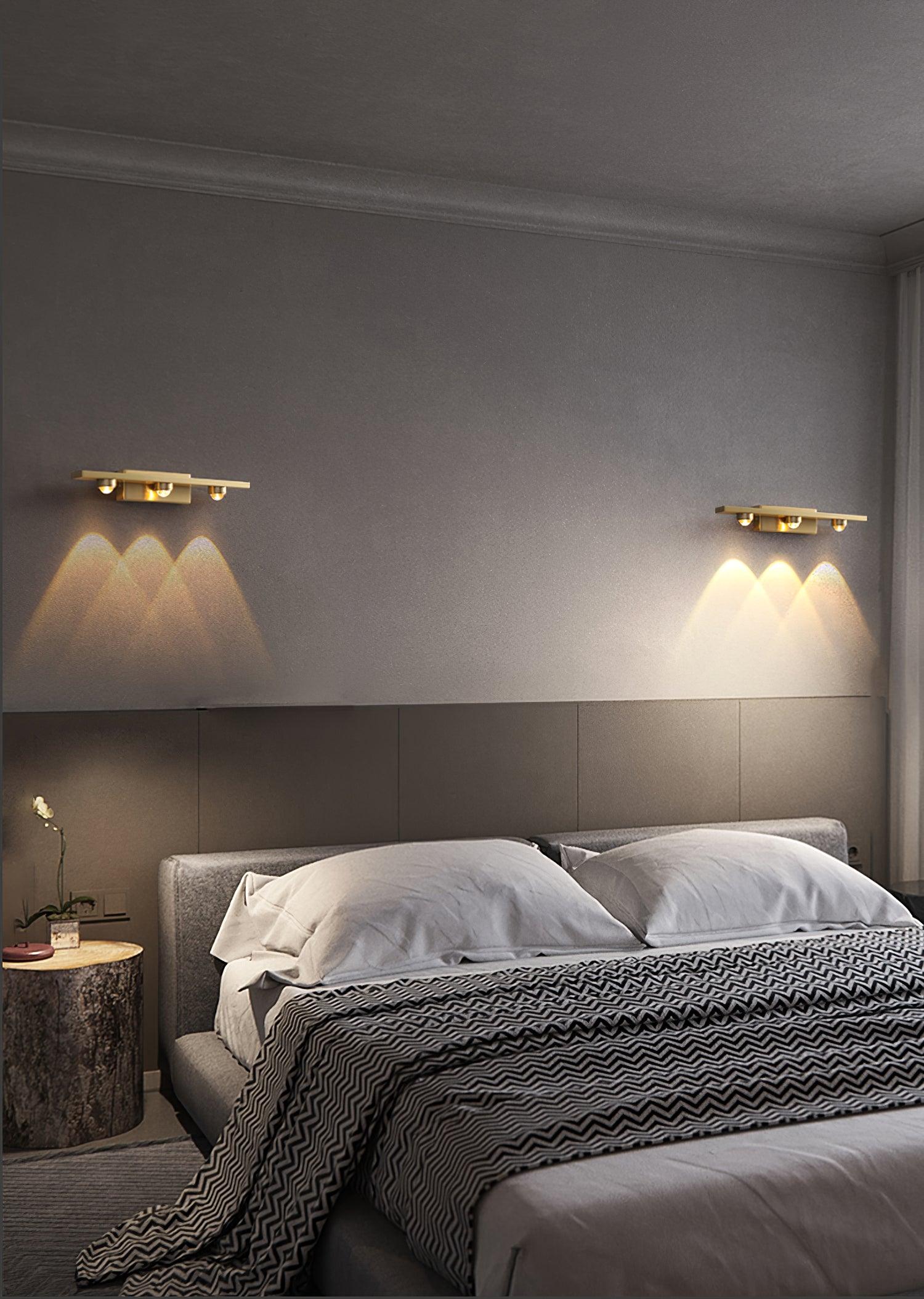 Wide bedroom view with two three-lens brass-tone wall lights flanking a grey upholstered headboard