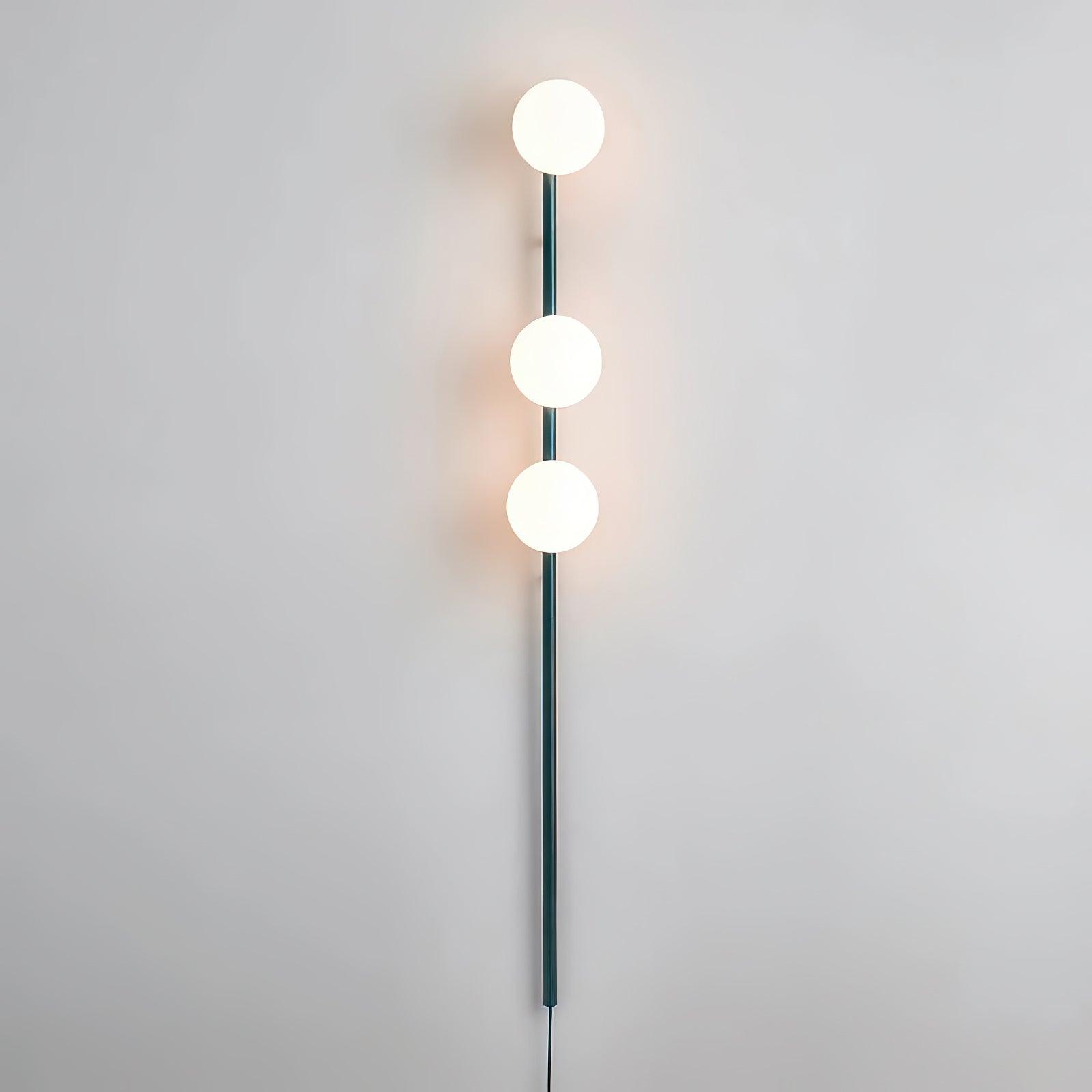 Lit teal wall light on pale wall, three glowing white globes along slim vertical rod and warm surrounding halos