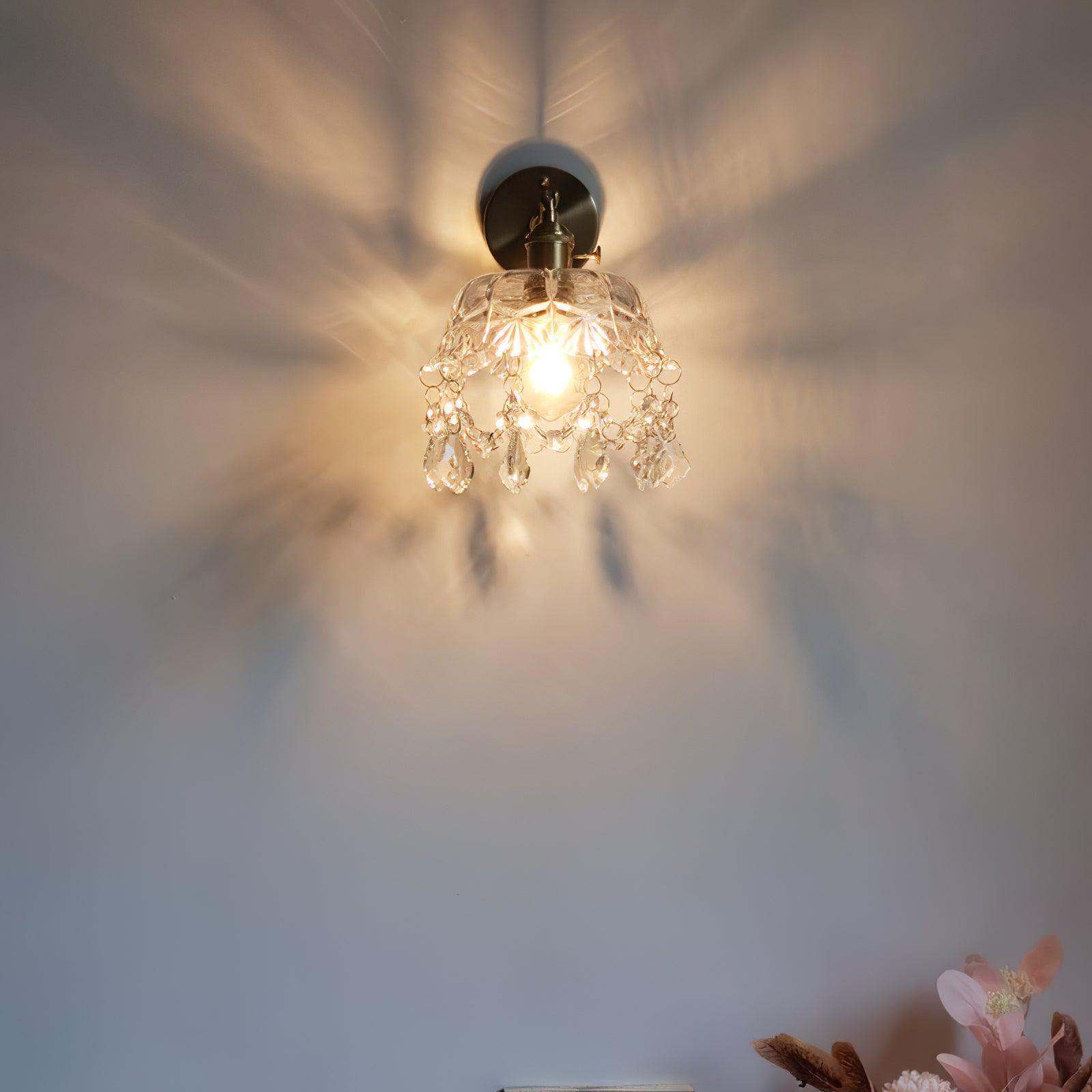 Farther front view of the lit bell-and-drop wall light, with patterned light on the wall and flowers below